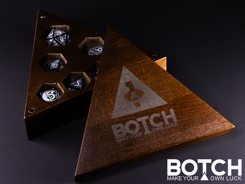 A triangular wooden dice box, the lid is laser engraved with the Botch logo and botch submark. A set of dice are shown in the box for an example fit, but do not come with it. The lid is held on by magnets. 